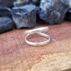 Puede incluir: Un anillo de plata con un diseño abierto único. El anillo presenta una banda lisa y redondeada que rodea el dedo, con una segunda banda que se curva por encima. El anillo se muestra sobre una superficie de madera, con rocas de color gris oscuro en el fondo.