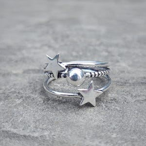 Puede incluir: Un conjunto de anillos plateados. Un anillo presenta una pequeña estrella, otro tiene un diseño de cuentas y un tercer anillo tiene una pequeña esfera. Los anillos están apilados, creando un efecto en capas. Los anillos están sobre una superficie gris.