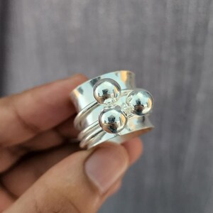 Può includere: Un anello spinner color argento con tre piccole perle rotonde e riflettenti. L'anello ha una fascia larga con un design strutturato. Le perle sono posizionate sulla parte superiore dell'anello, aggiungendo un dettaglio unico al gioiello.