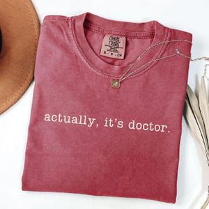 Puede incluir: Una camiseta Comfort Colors color burdeos con el texto "actually, it's doctor." Un collar plateado con un pequeño colgante está sobre la camiseta doblada. El fondo es de color claro.