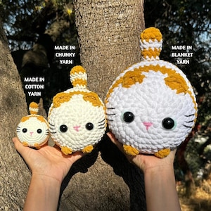 May include: Three crocheted cat toys in varying sizes, held against a tree trunk. The cats are white with orange markings, black whiskers, and large black eyes. Text indicates they are made with cotton, chunky, and blanket yarn.