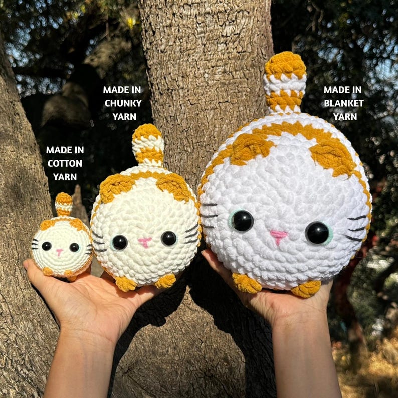 May include: Three crocheted cat toys in varying sizes, held against a tree trunk. The cats are white with orange markings, black whiskers, and large black eyes. Text indicates they are made with cotton, chunky, and blanket yarn.