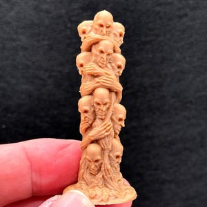 Może przedstawiać: Szczegółowa, brązowa rzeźba przedstawiająca wieżę z czaszek. Czaszki są ułożone jedna na drugiej, niektóre wydają się obejmować. Rzeźba ma teksturowaną podstawę i jest trzymana przez dłoń na ciemnym tle.