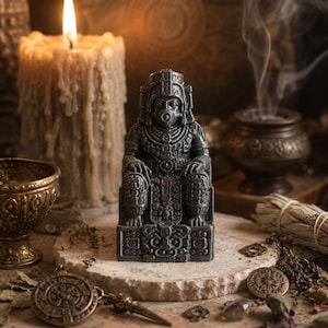 Può includere: Una statuetta grigio scuro con finitura metallica che raffigura una divinità seduta con intricati motivi geometrici intagliati e un copricapo. La figura è in posizione centrale ed eretta, mostrando dettagliati lavori in rilievo sul busto.