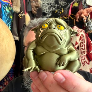 Puede incluir: Una pequeña figura metálica de color verde oliva de una criatura robusta y triste con grandes ojos amarillos llorosos, orejas puntiagudas y boca hacia abajo. La figura tiene un cuerpo liso y redondeado con extremidades visibles.