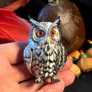 Könnte beinhalten: Silberfarbene Eulenfigur mit orangefarbenen Augen und gelbem Schnabel. Die Eule hat detaillierte Federmuster und wird in einer Hand gehalten. Die Figur ist ein dekoratives Objekt.