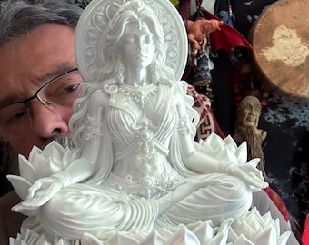 Puede incluir: Una figura blanca detallada de una deidad sentada en posición de meditación con las piernas cruzadas sobre una base de flor de loto grande de múltiples pétalos. La figura presenta un halo circular ornamentado y joyas intrincadas.