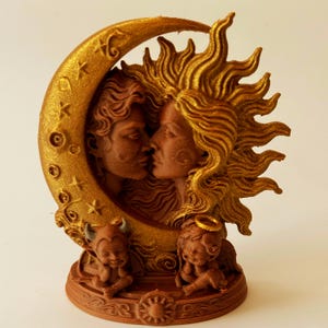 Può includere: Una scultura in metallo dorato e marrone raffigurante una luna crescente e un sole con volti umani di profilo, posizionati l'uno di fronte all'altro. Sotto, una piccola figura cornuta e una figura alata con aureola siedono su una base circolare.