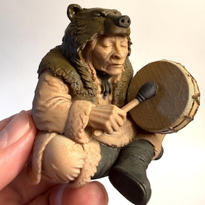 May include: A detailed figurine of a person wearing a bear headdress and a fur-lined coat, holding a drum and drumstick. The figure is in a seated position, with closed eyes, and is rendered in shades of brown and green.