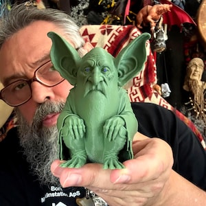 May include: A small, matte green figurine of a creature with large, pointed ears, a wrinkled face, and a long, flowing beard. The creature is depicted in a seated, hunched posture with its hands resting on its knees, featuring detailed facial textures.