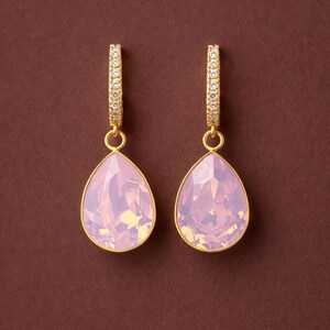 May include: A pair of gold-toned hoop earrings with teardrop-shaped, light pink crystal pendants. The hoops are encrusted with small, clear crystals. The earrings hang against a brown background.