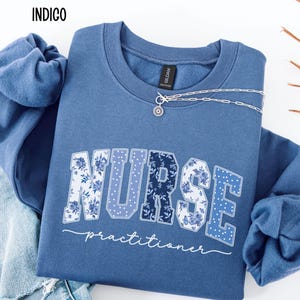 May include: Indigo blue sweatshirt with the word "NURSE" in a floral design and the word "practitioner" in white script. A silver necklace with a small pendant is draped on the sweatshirt. The word "INDIGO" is in the upper left corner.