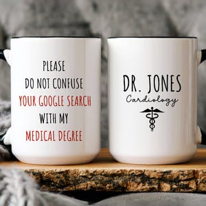 May include: Two white ceramic mugs with black handles and rims. One mug reads "PLEASE DO NOT CONFUSE YOUR GOOGLE SEARCH WITH MY MEDICAL DEGREE" in black and red text. The other mug says "DR. JONES Cardiology" with a medical symbol.