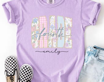 May include: A light purple short-sleeve t-shirt featuring the word GRADE in block letters with floral, gingham, and striped patterns, overlaid with the cursive text fourth and the name emily. The shirt is paired with light blue denim shorts.
