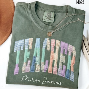 May include: A moss green t-shirt with the word "TEACHER" in patchwork letters. The shirt also features the name "Mrs. Jones" in script. A gold necklace with a pendant rests on the shirt.