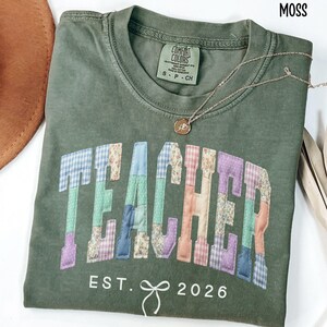 May include: A moss green t-shirt with the word "TEACHER" in colorful patchwork letters. The text "EST. 2026" is below the word "TEACHER". A gold necklace with a pendant is draped across the shirt. A brown hat is visible in the upper left corner.
