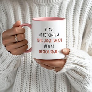 White ceramic mug with a pink handle and interior, featuring 'PLEASE DO NOT CONFUSE YOUR GOOGLE SEARCH WITH MY MEDICAL DEGREE' in red and black.