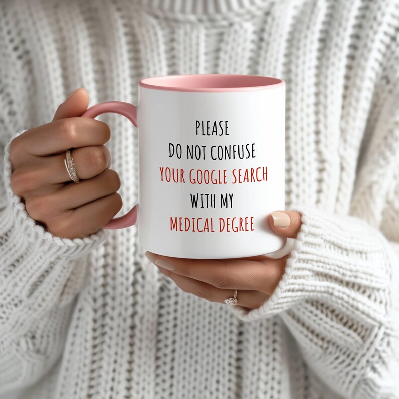 White ceramic mug with a pink handle and interior, featuring 'PLEASE DO NOT CONFUSE YOUR GOOGLE SEARCH WITH MY MEDICAL DEGREE' in red and black.
