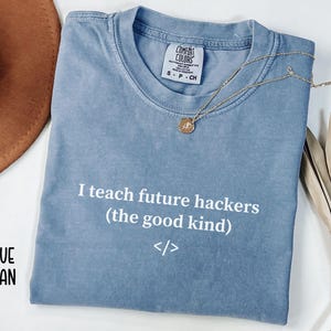 May include: A blue jean-colored t-shirt with the text "I teach future hackers (the good kind)". A gold necklace and a brown hat are also in the image. The shirt is made by Comfort Colors.