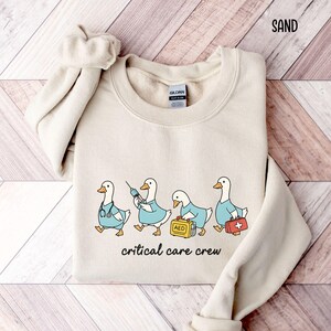 A "Sand" sweatshirt, featuring four cartoon geese dressed as an ICU medical team, with the text "critical care crew" printed below.