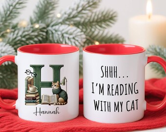 May include: Two white ceramic mugs with red interiors and handles. One mug features a green letter "H" with a cat, books, and the name "Hannah." The other mug says "SHHH... I'M READING WITH MY CAT."