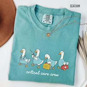 A "Seafoam" Comfort Colors t-shirt featuring four cartoon geese dressed as an ICU medical team, with the text "critical care crew" printed below.