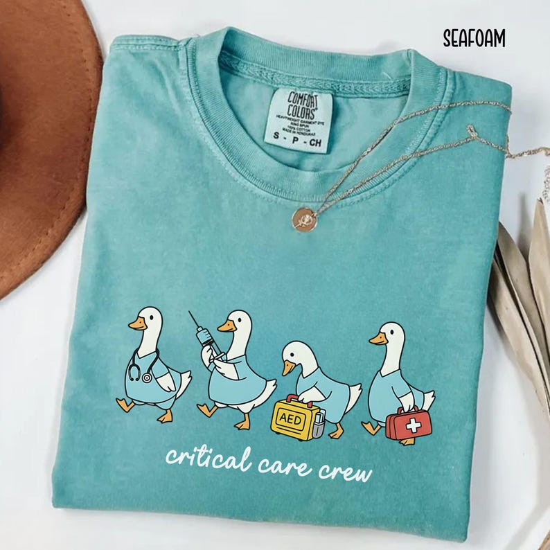 A "Seafoam" Comfort Colors t-shirt featuring four cartoon geese dressed as an ICU medical team, with the text "critical care crew" printed below.