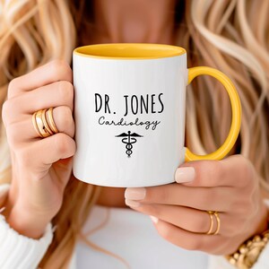 White ceramic mug with a yellow rim and handle. Features a custom personalized design with 'Dr. Jones Cardiology' and a medical symbol.