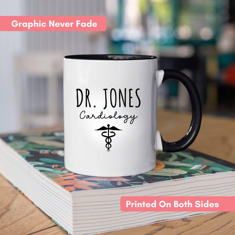 White ceramic mug with a black rim and handle. Features a custom personalized design with 'Dr. Jones Cardiology' and a medical symbol. Includes pink banners highlighting key features: 'Graphic Never Fade' and 'Printed On Both Sides'."