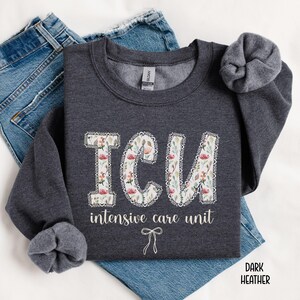 A "Dark Heather" sweatshirt, featuring the letters "ICU" in a block font filled with a green, pink, and white floral pattern with a decorative border, and the words "intensive care unit" and a bow below.