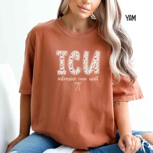A "Yam" Comfort Colors t-shirt, featuring the letters "ICU" in a block font filled with a green, pink, and white floral pattern with a decorative border, and the words "intensive care unit" and a bow below.