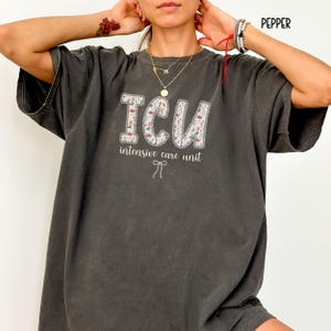 A "Pepper" Comfort Colors t-shirt, featuring the letters "ICU" in a block font filled with a green, pink, and white floral pattern with a decorative border, and the words "intensive care unit" and a bow below.