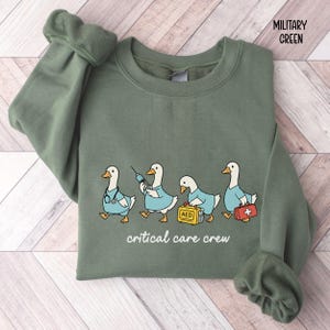 A "Military Green" sweatshirt, featuring four cartoon geese dressed as an ICU medical team, with the text "critical care crew" printed below.