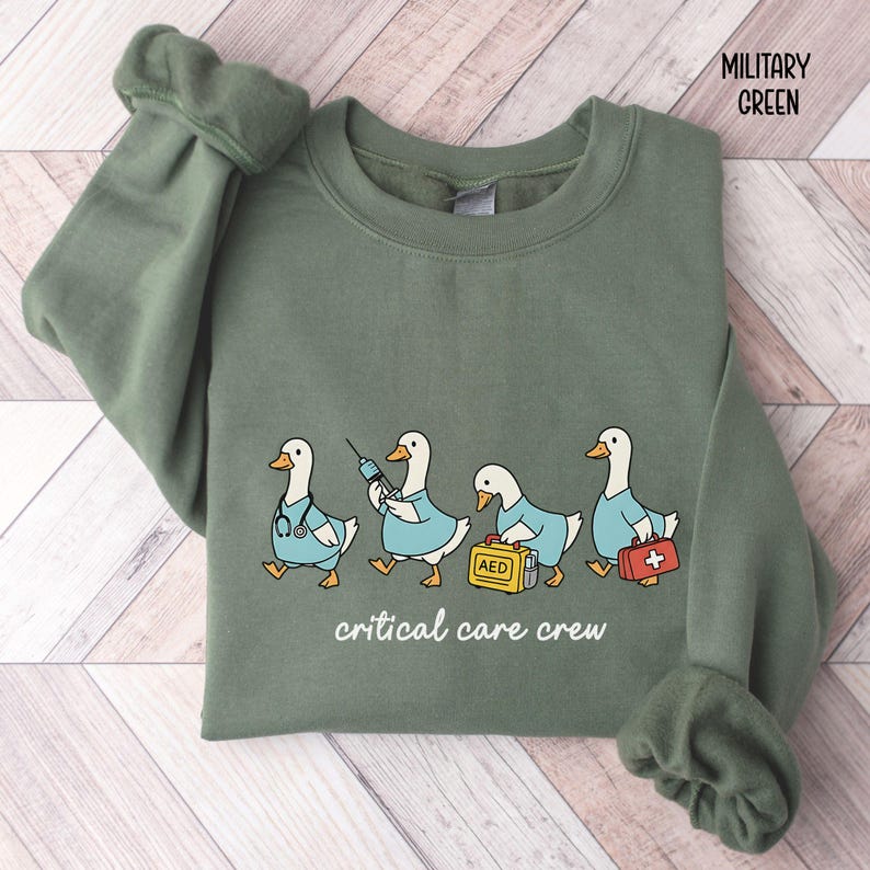 A "Military Green" sweatshirt, featuring four cartoon geese dressed as an ICU medical team, with the text "critical care crew" printed below.