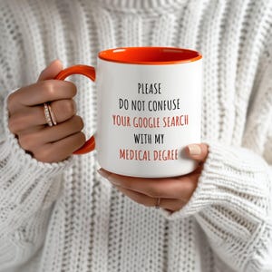 White ceramic mug with a orange handle and interior, featuring 'PLEASE DO NOT CONFUSE YOUR GOOGLE SEARCH WITH MY MEDICAL DEGREE' in red and black.