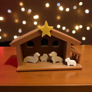 May include: A brown miniature nativity scene with a yellow star on top. The scene includes four dachshund dog figurines in shades of white and gray, inside a small wooden structure. The background features blurred lights.