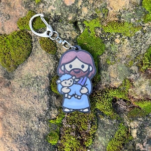 May include: A cartoon keychain featuring a figure with brown hair and a beard, dressed in blue robes, holding a blue lamb. The keychain has a silver clasp. The background is a stone surface covered in green moss.
