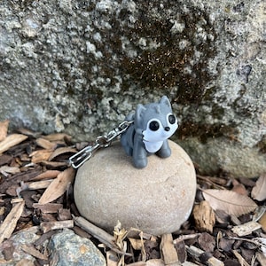 May include: A gray wolf keychain with a silver chain. The wolf figurine has black eyes and white accents. It is sitting on a smooth, light brown stone, surrounded by wood chips and leaves, against a textured rock background.