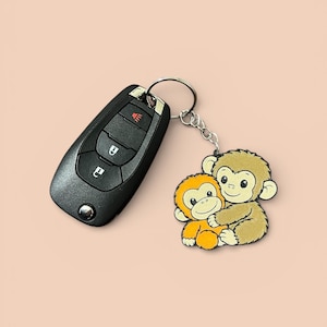 May include: A black car key next to a keychain with a cartoon image of two monkeys hugging. One monkey is orange, and the other is tan. The keychain is attached to a silver metal ring.