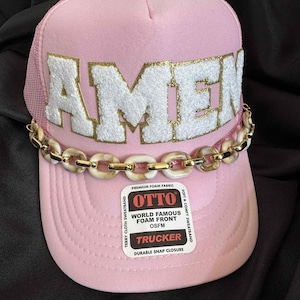 May include: Pink trucker hat with the word "AMEN" in white, outlined in gold, across the front. A gold and cream chain is attached to the brim. The hat has a mesh back and a "OTTO" label.