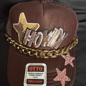 Puede incluir: Una gorra de camionero marrón con una estrella grande de lentejuelas doradas y la palabra HOWDY en letras de lentejuelas. Una cadena dorada gruesa cruza la parte frontal. Dos estrellas de lentejuelas rosas más pequeñas decoran la visera.