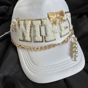 Peut inclure: Casquette de baseball blanche avec le mot "WIFEY" en or et blanc, ornée d'un nœud et d'une chaîne dorés, ainsi que de perles. La casquette a un dos en filet et une visière incurvée.