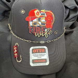 May include: A black trucker hat with a mesh back and a foam front. The hat features an embroidered design of a firefighter's gear and the words "FIRE WIFE". It is accessorized with a gold chain and a red beaded pin.