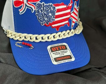 May include: A blue trucker hat with a white mesh back and a cream-colored chain across the brim. The front features patriotic patches including a bow, a map of the USA, and a star. The hat has the text "OTTO TRUCKER" on the front.