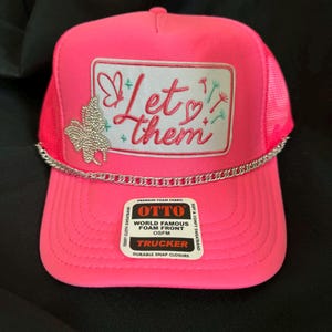 May include: A bright pink trucker hat with a mesh back and a curved brim. The front features a rectangular patch with the words "Let them" in cursive, along with butterfly and floral designs. A silver chain accents the brim. The hat has a snap closure.