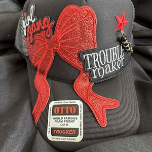 May include: Black trucker hat with a red glitter bow and the words "girl gang" and "trouble maker". The hat has a mesh back and a snap closure. A red star charm hangs from the side.