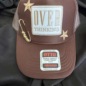 May include: Brown trucker hat with a mesh back and a curved brim. The front features the text "OVER THINKING" in gold lettering on a white patch, along with two gold star accents and a gold safety pin. The hat has a snap closure.