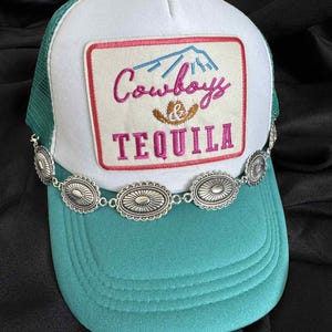 Puede incluir: Gorra de camionero con panel frontal blanco, visera turquesa y parte trasera de malla verde. La gorra presenta un parche con las palabras "Cowboys TEQUILA" en rosa y azul, y una cadena plateada con adornos ovalados.