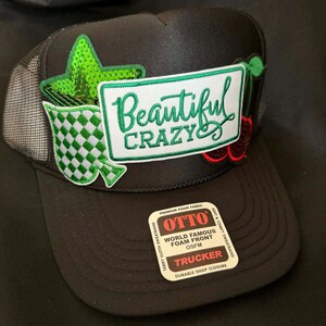 Puede incluir: Gorra de camionero negra con parte trasera de malla y visera curva. La parte delantera presenta un parche con las palabras "Beautiful Crazy" en verde, junto con parches de estrella y cereza con lentejuelas verdes. Cierre a presión.