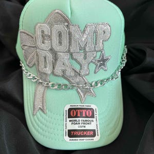 May include: A mint green trucker hat with the text "COMP DAY" in silver sequined letters. A silver chain is draped across the front. The hat has a curved brim and a mesh back. The hat has a "OTTO" label.